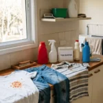 Uma bancada de uma lavanderia doméstica com roupas sujas e produtos para usar como tirar manchas de base na roupa.